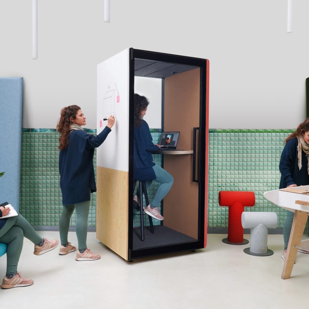 Afbeelding van een kantoor met verschillende werkplekken. Je ziet een kleine werkpod waar iemand in een online meeting zit. Aan de buitenkant is een whiteboard waar iemand opschrijft.