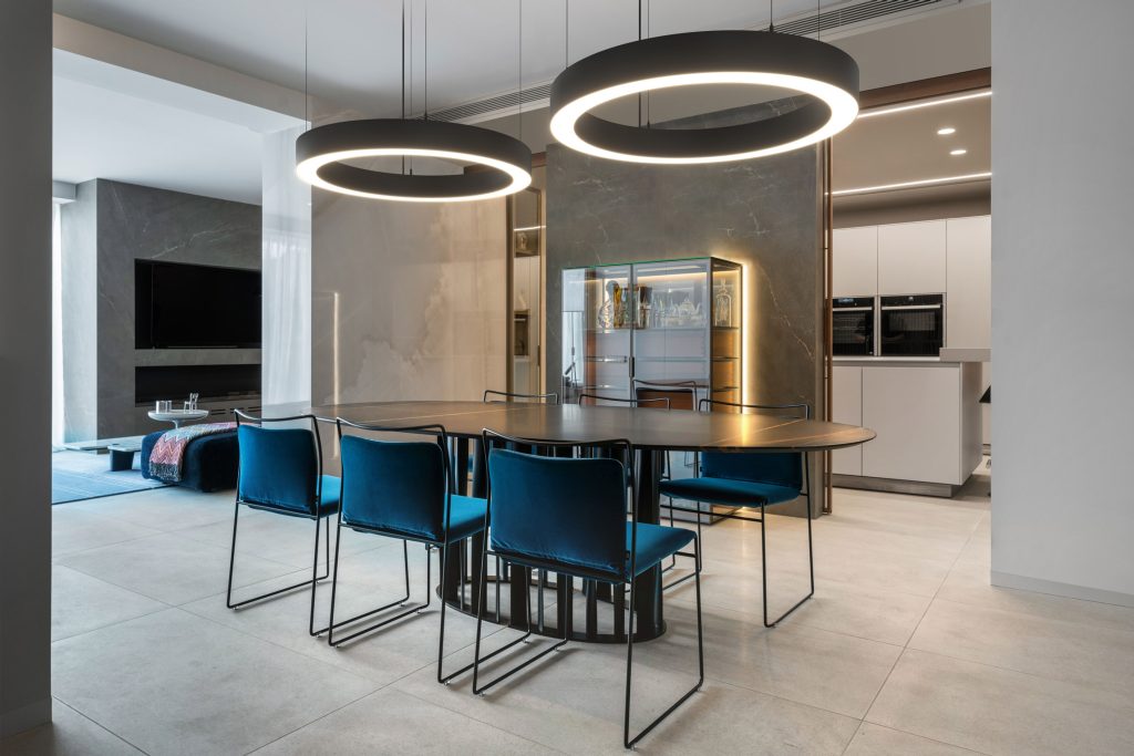 Semi-open keuken met ovale eettafel en blauwe stoelen. Boven de tafel hangen twee ronde lampen 'Glorious van PROLICHT'.