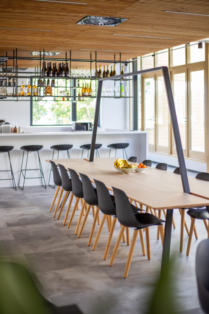 Zijaanzicht van een bar met daarboven rekken met drank, een wit keukenblad met stoelen en daarvoor een lange eettafel van Lande.