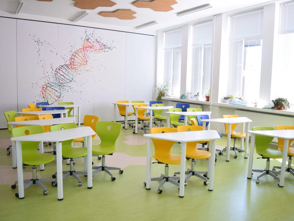 Klaslokaal met dna print op de muur met daarvoor witte bureaus en gele, blauwe of groene stoelen.