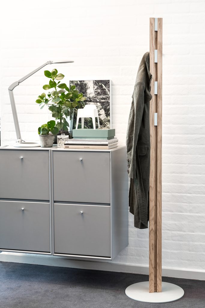 Grijze kast met vier vakjes, lamp, plant en afbeelding naast een kapstop met jas.